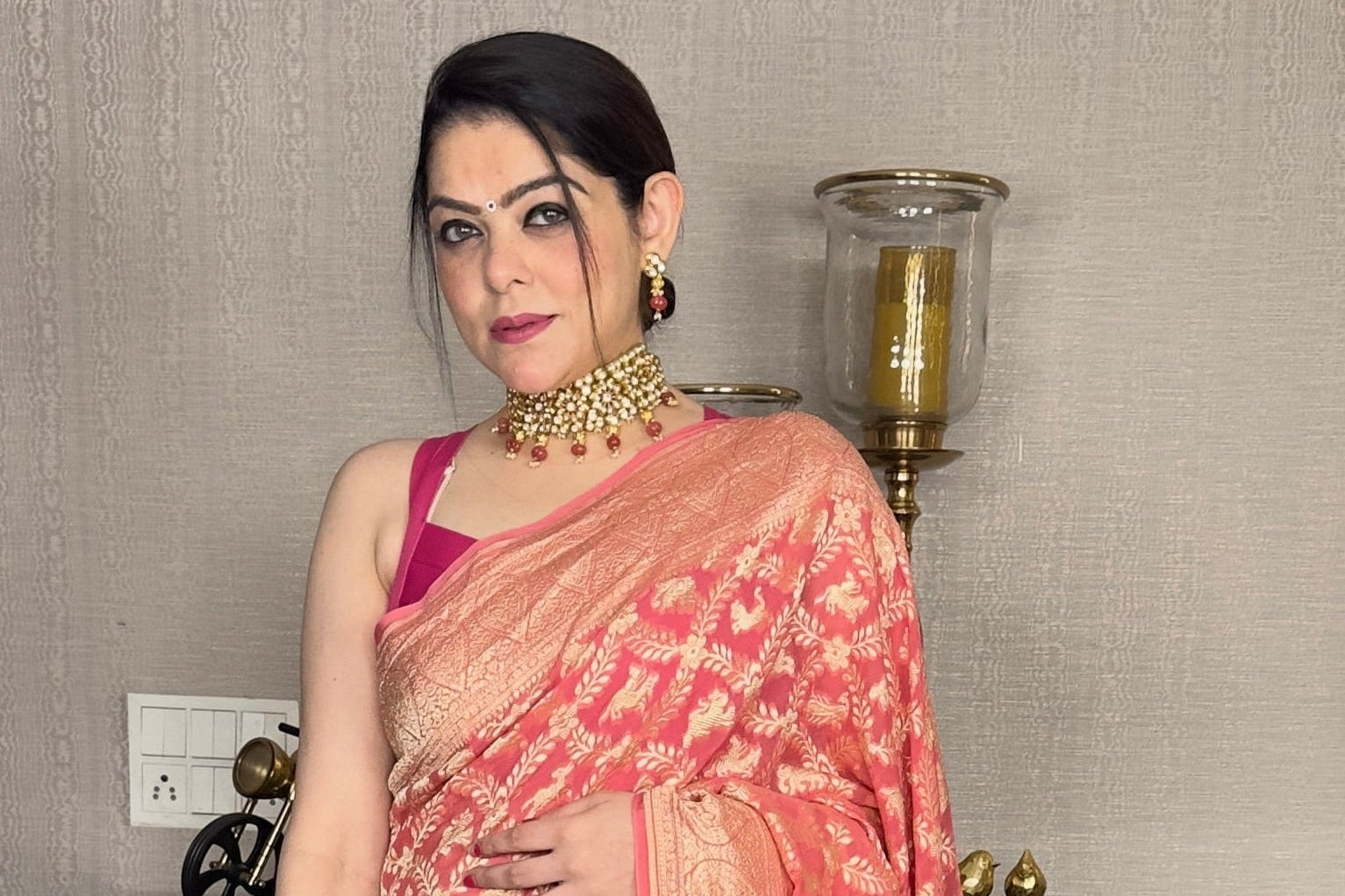 Woman in a pink saree standing indoors with decorative elements in the background